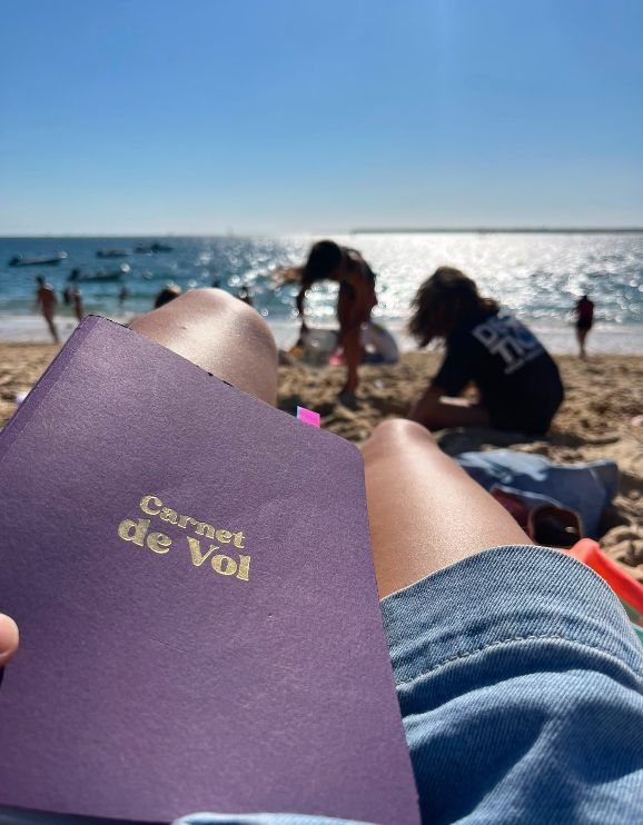 Le Carnet de Vol du Village, posé sur les genoux d'une Villageoise à la plage